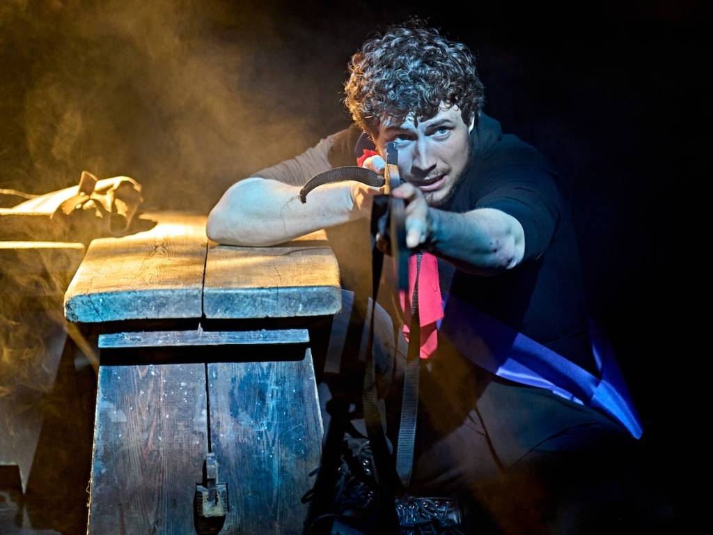 un uomo nella produzione teatrale *Martin Guerre* sembra tenere in mano un oggetto di scena simile a una pistola mentre si sporge da una panchina di legno