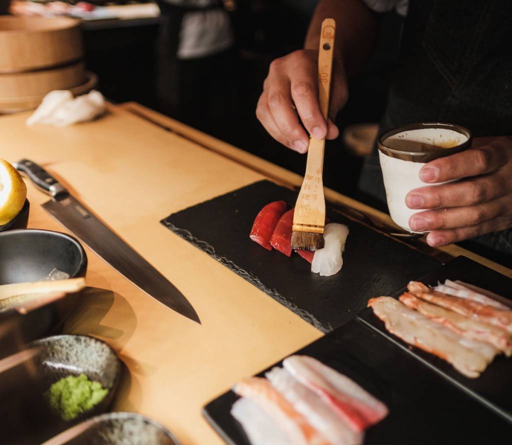 A person is brushing sauce onto sushi, with more food prepped on cutting boards