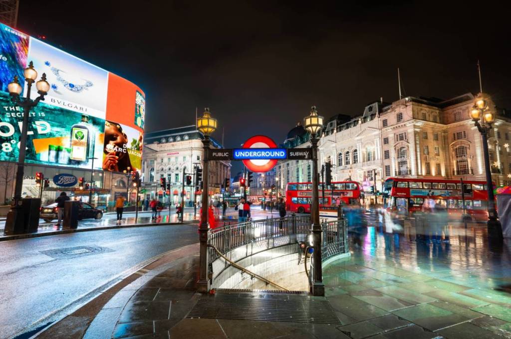 Piccadilly Circus à noite, com o letreiro da entrada do metro no centro e os ecrãs e a estrada à sua volta.