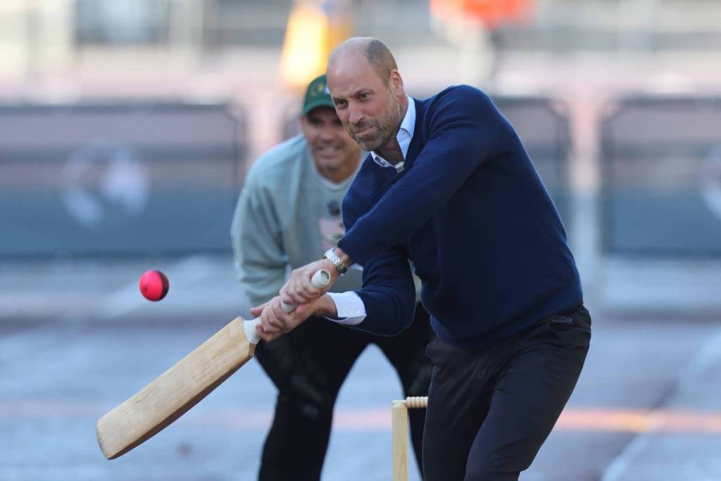 El príncipe Guillermo sostiene un bate y una pelota roja viene hacia él; va vestido de azul marino y se ven las puertas del Palacio de Buckingham a lo lejos