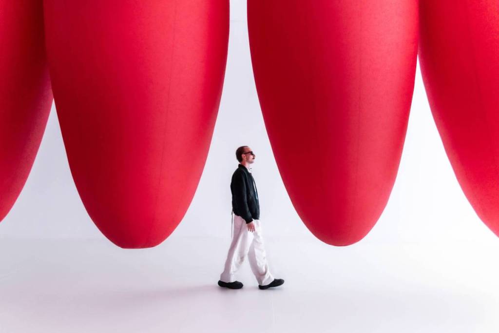 Un uomo che cammina sotto un’installazione artistica sospesa composta da grandi ovoidi rossi in una stanza altrimenti completamente bianca: una delle installazioni artistiche dell’esperienza Euphoяia al Balloon Museum