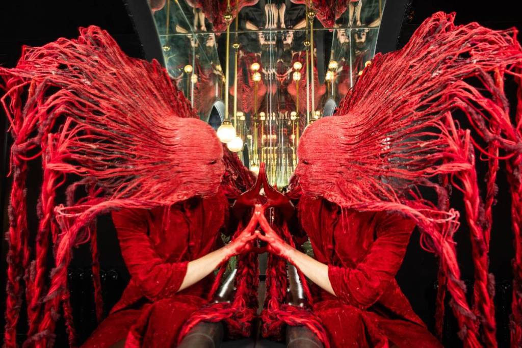 A person wearing a textured red mask and red clothing stands in front of a mirror, pressing their fingers against their reflection.