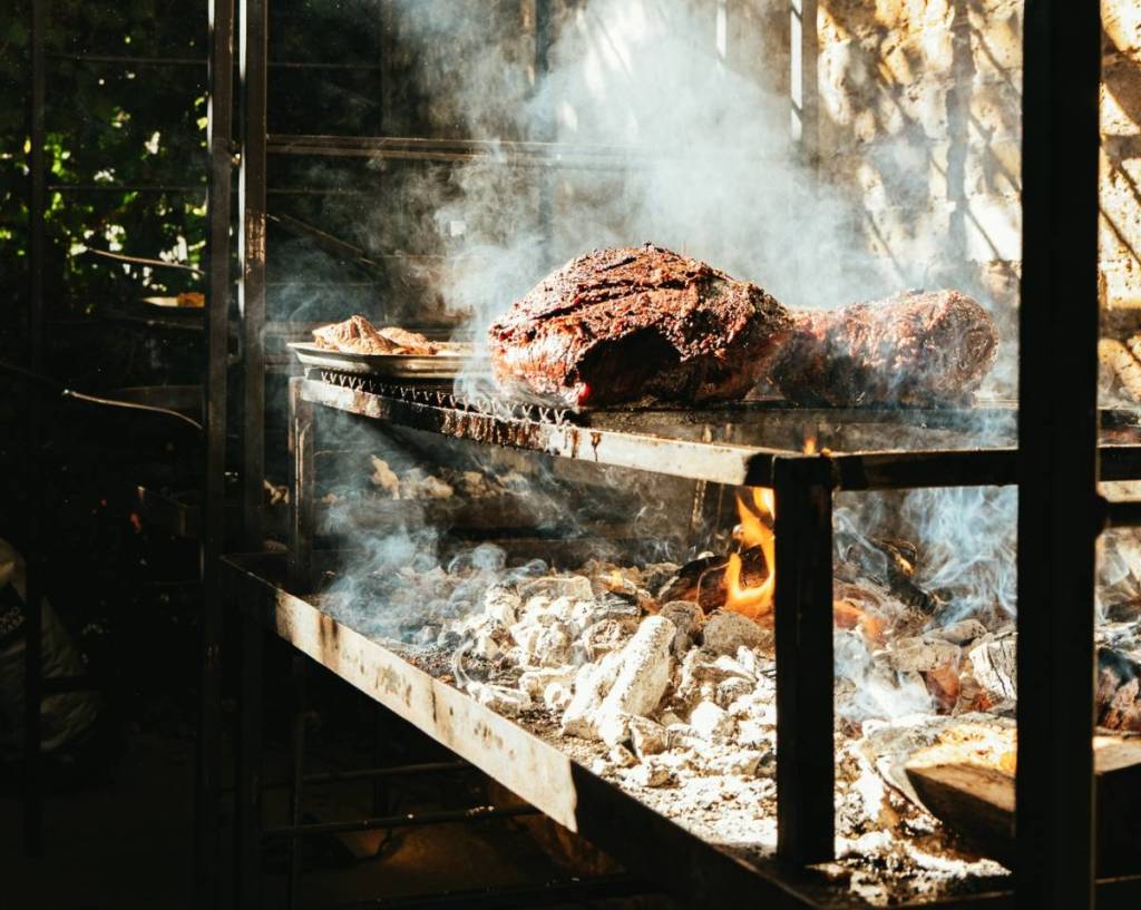 Large cuts of meat cooking on a metal grill over hot coals.
