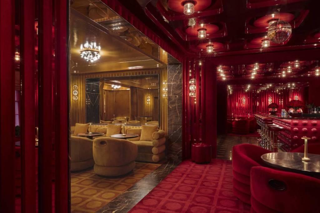 A lavish Art Deco lounge glows in red and gold, with velvet seating, mirrored walls, chandeliers, and a bar