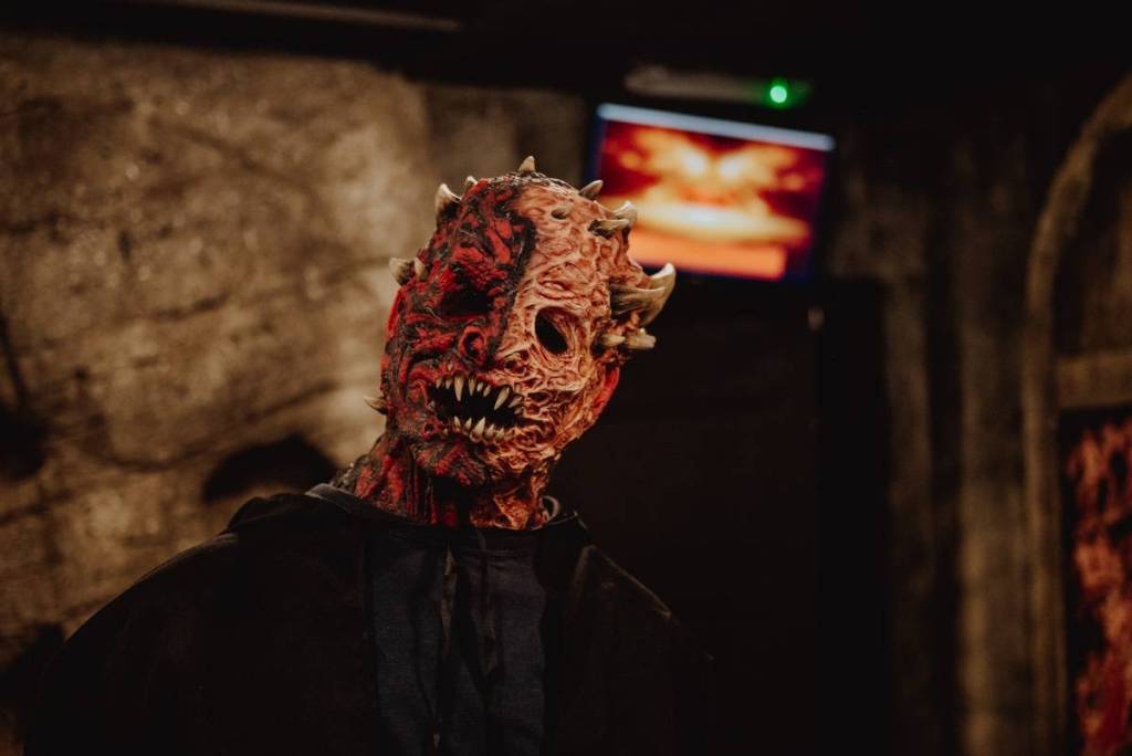 A figure in black wears a grotesque red-and-tan demon mask with horns and sharp teeth in a dim stone room
