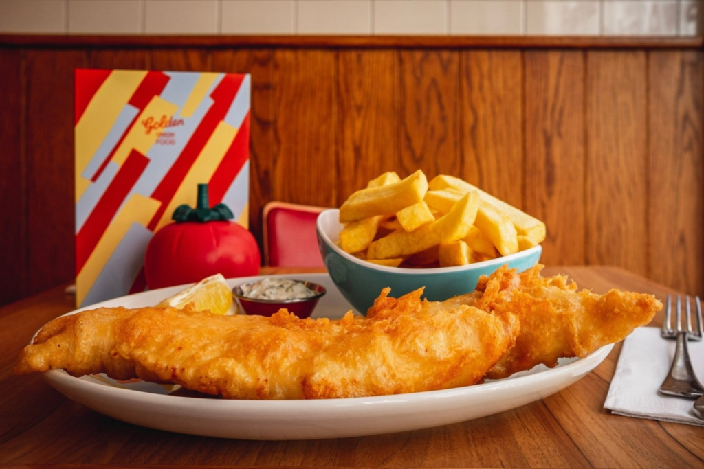 Un piatto di fish and chips su un tavolo con un menu e un dispenser di ketchup a forma di pomodoro