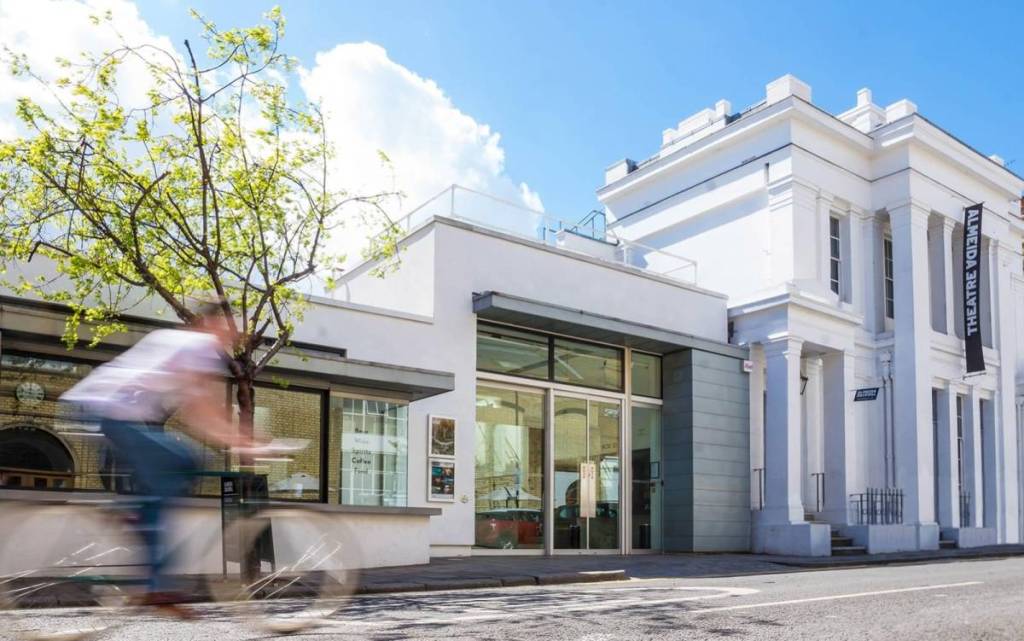Jemand auf einem Fahrrad, der an einem sonnigen Tag am weißen Gebäude des Almeida Theatre vorbeifährt