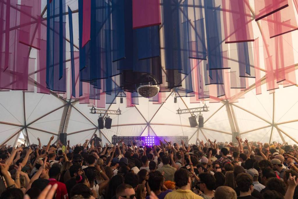 Una folla festante alza le mani sotto pannelli sospesi traslucidi rossi e blu, con un palco per i DJ che risplende di viola in un tendone luminoso