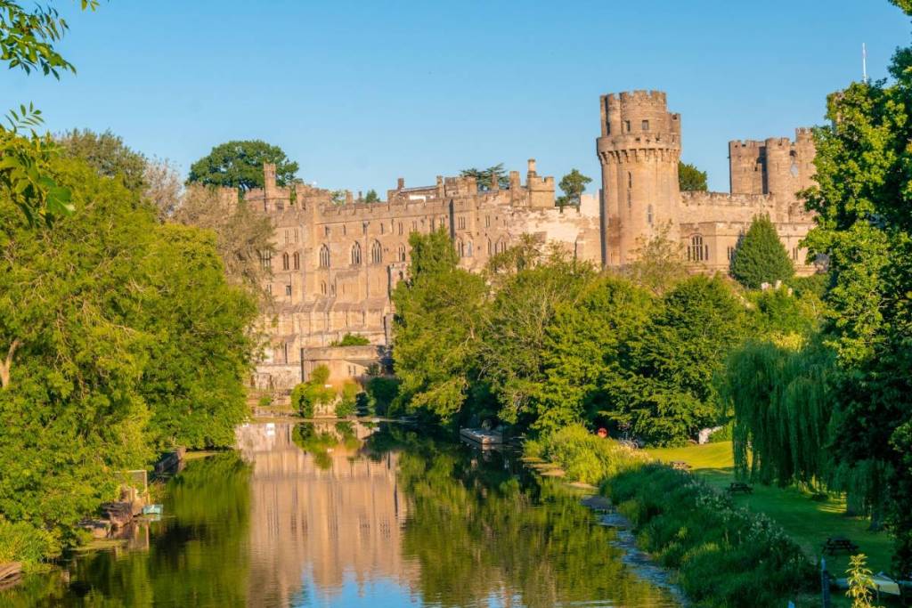 el exterior del castillo de Warwick, rodeado de árboles frondosos y un estanque en un día soleado
