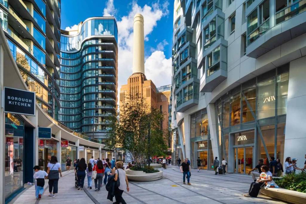 persone che passeggiano e si siedono sulle panchine lungo un sentiero nel complesso della Battersea Power Station, con ristoranti e negozi che fiancheggiano il percorso e altri edifici visibili in alto, oltre a una delle iconiche ciminiere della Battersea Power Station