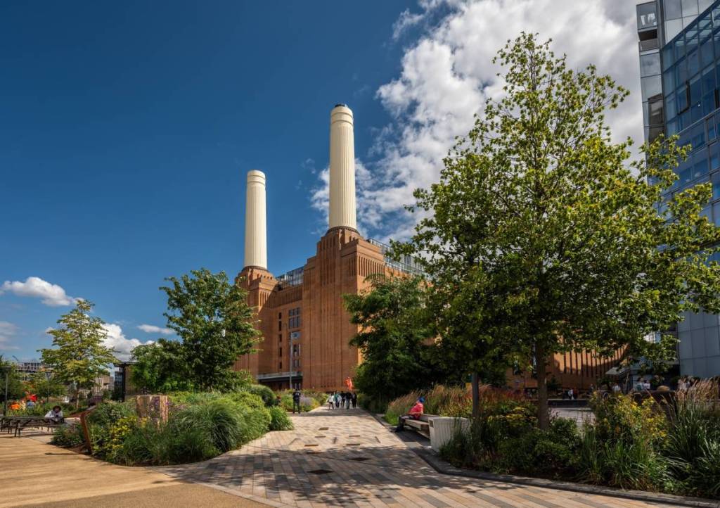 La Battersea Power Station vista dal parco a nord dell’edificio, incorniciata da alberi e fogliame, in una giornata di cielo azzurro