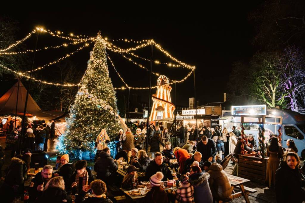 una vista notturna di un mercatino di Natale con un grande albero di Natale, giostre e gente seduta ai tavoli a mangiare e bere bevande calde