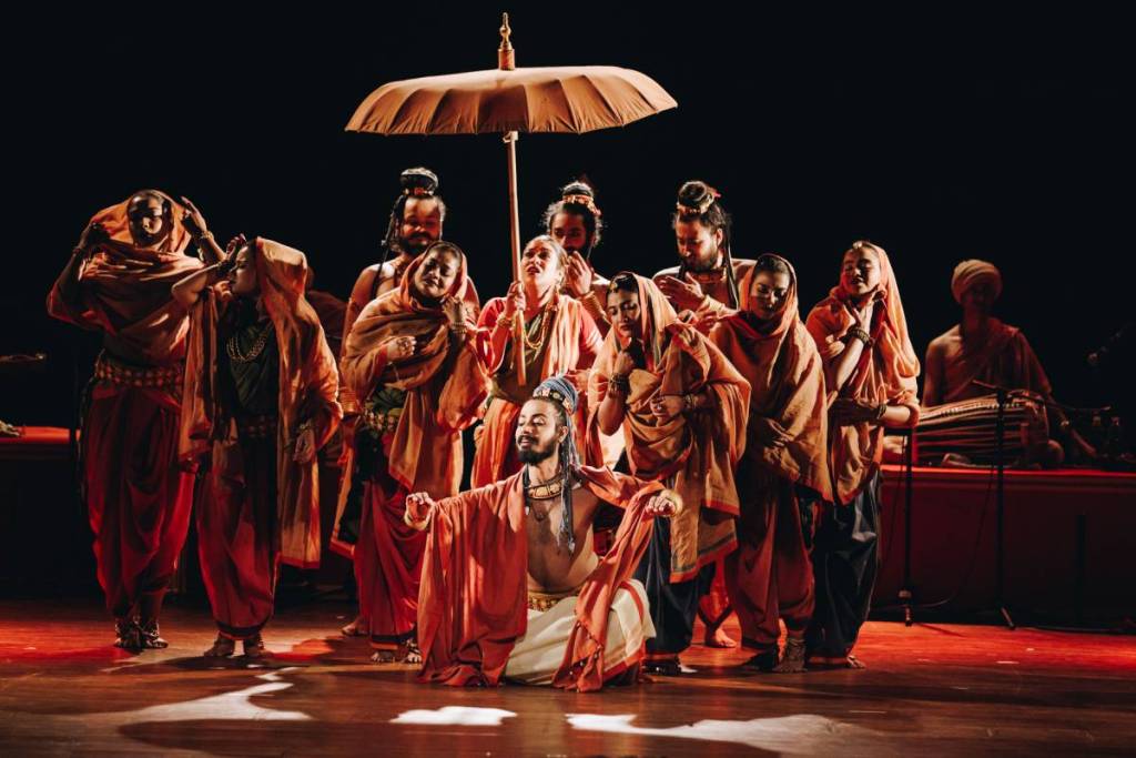 a group of performers on stage in traditional south asian garments, clustered around each other trying to get under a wooden umbrella