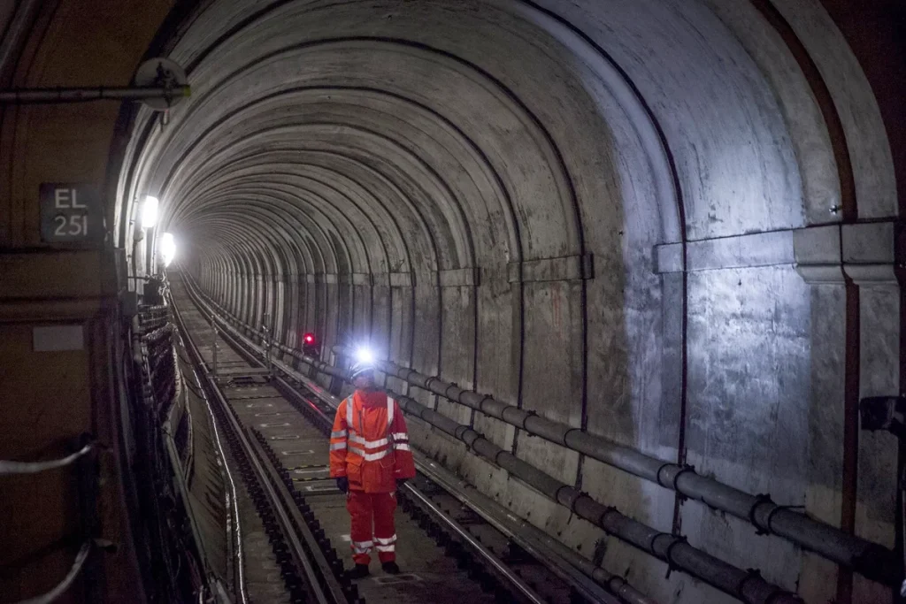 Um trabalhador ferroviário com equipamento de segurança laranja vivo está nos carris, dentro de um longo túnel de betão abobadado, iluminado por uma lanterna