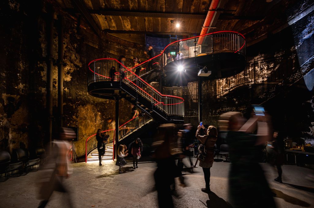 Uma escadaria em espiral vermelha serpenteia por um salão industrial escuro, enquanto visitantes difusos se movem lá em baixo, criando uma cena dramática e atmosférica no museu.