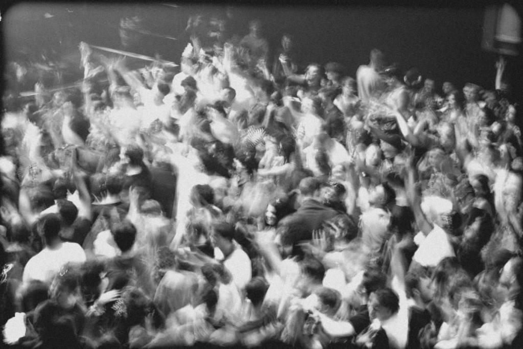 Grainy black-and-white photo of a densely packed crowd, with people raising their arms and surging together 