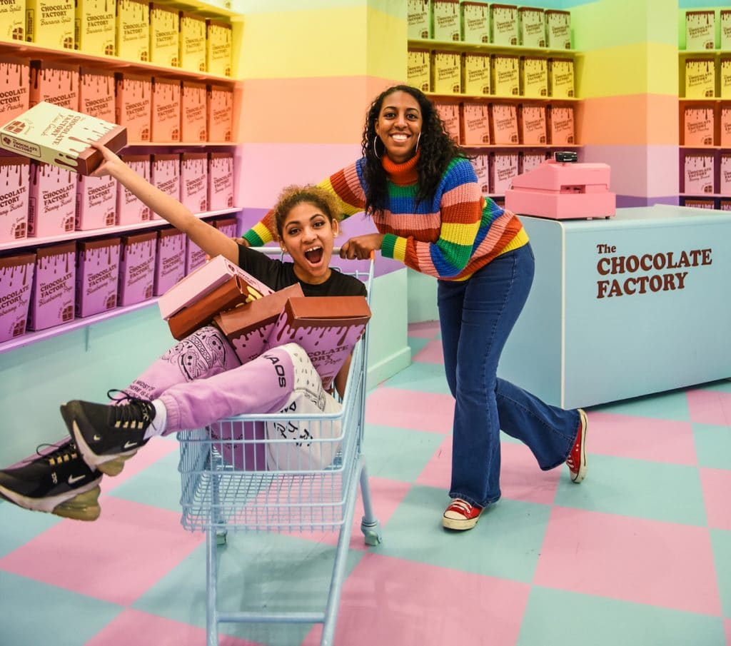 girl pushing another girl in a trolley surrounded by pastel-coloured cereal boxes