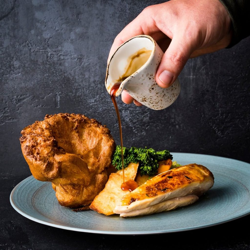 Somebody pouring gravy over a Sunday dinner, consisting of a huge Yorkshire pudding, broccoli and roast chicken at Barnacle.
