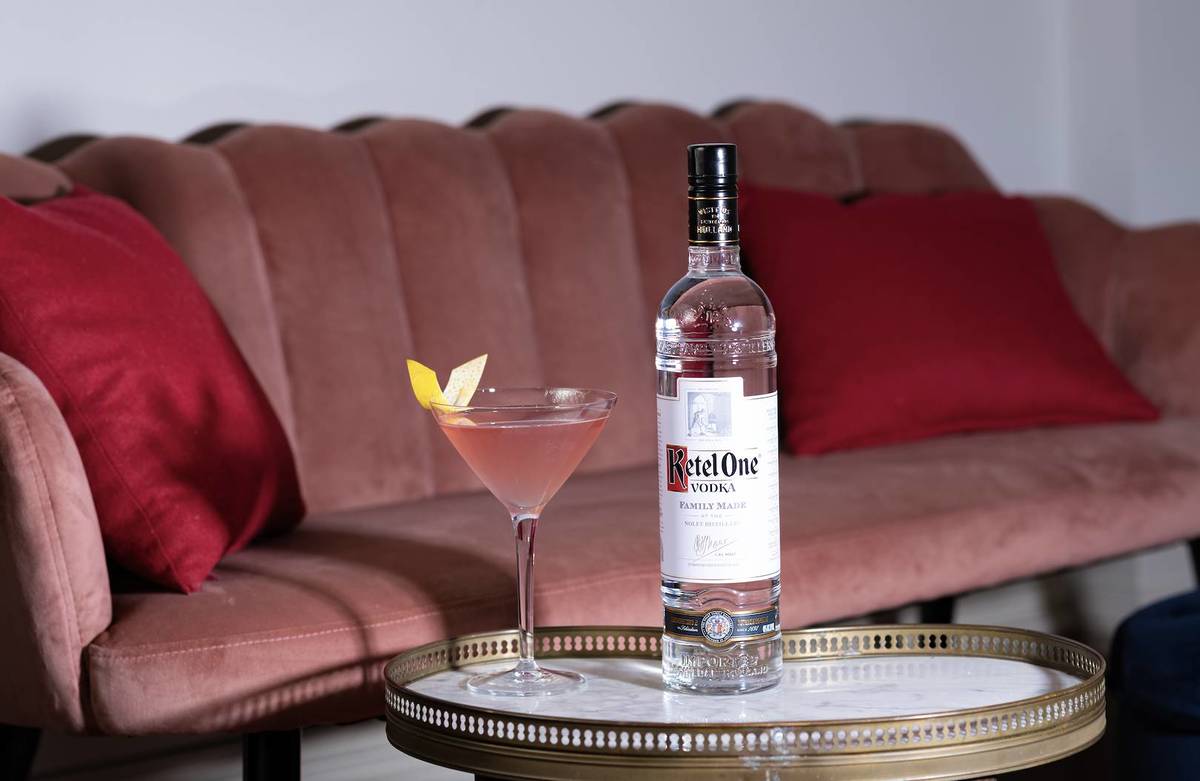 A cosmo next to a Ketel One Vodka bottle in front of a pink sofa.