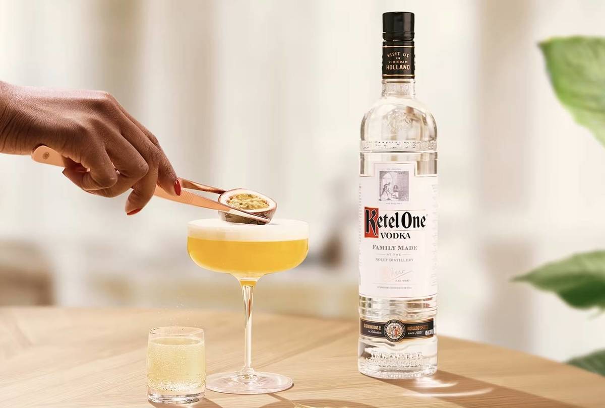 A hand is placing a passionfruit as garnish on a Passionfruit Martini next to a Ketel One Vodka bottle.