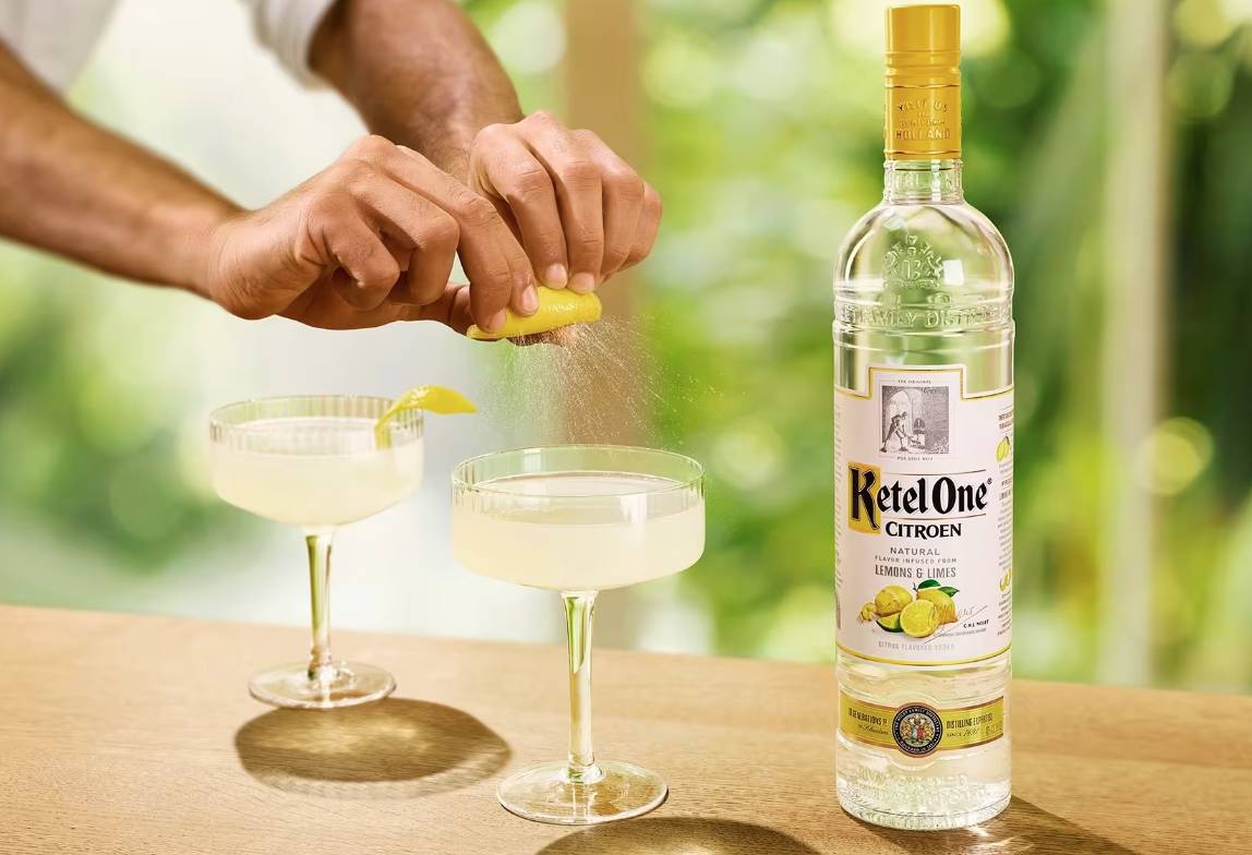 Hands squeeze a lemon into two glasses of Lemon Drop Martini sitting next to a bottle of Ketel One Citroen.