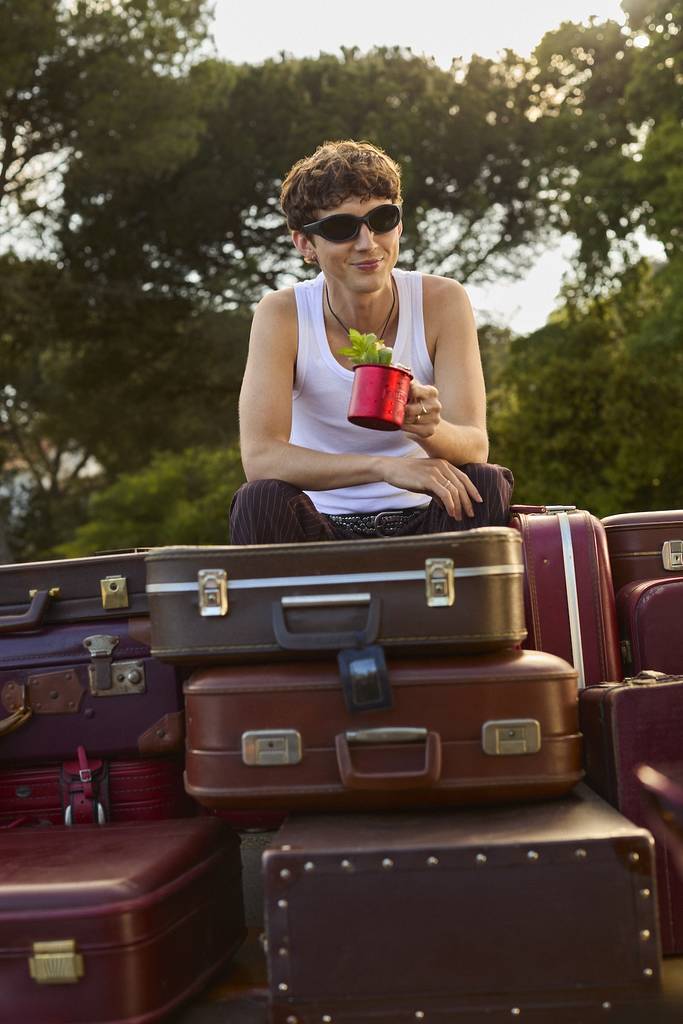 Troye sits on a pile of suitcases with a cocktail in a red cup in hand.