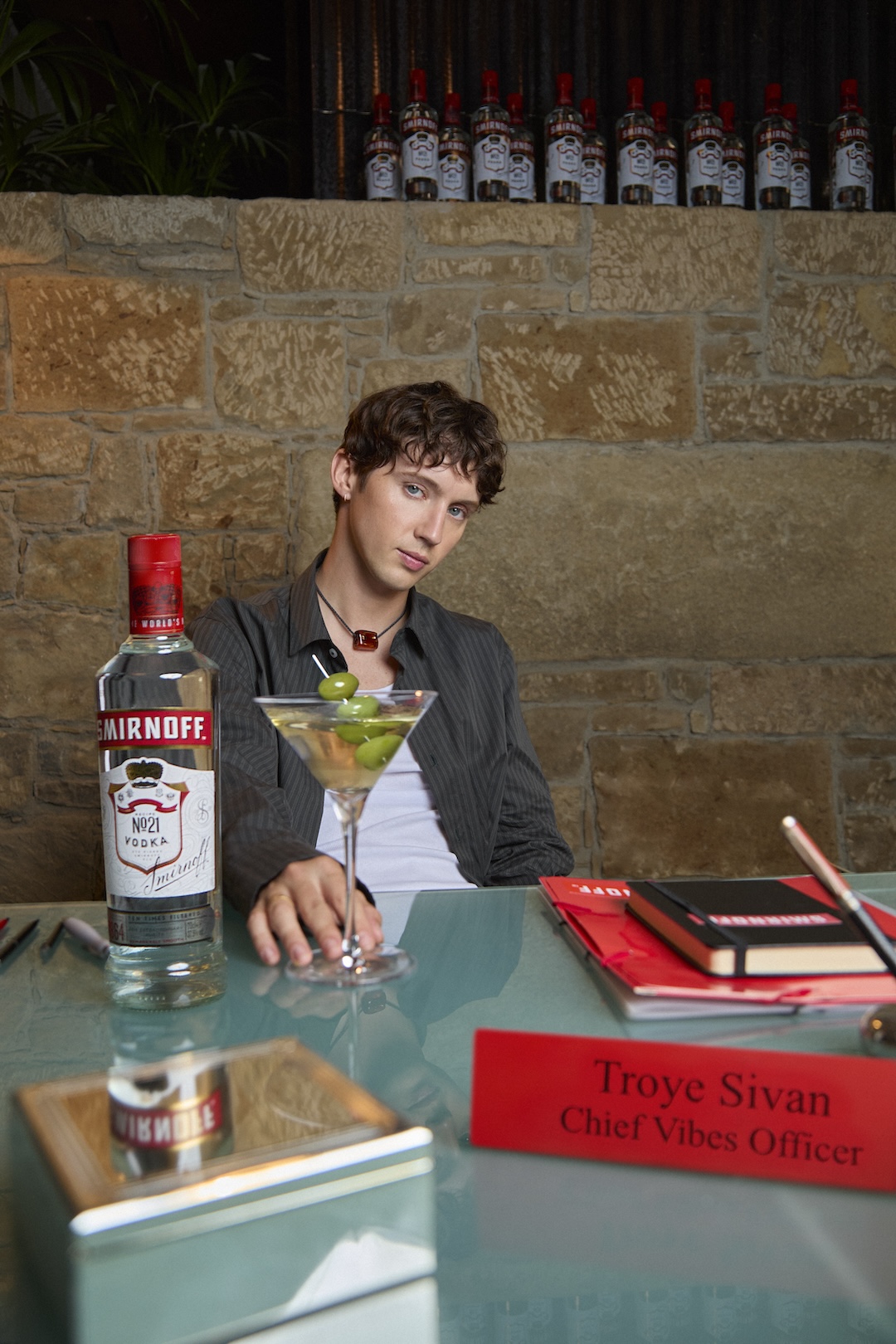 Troye Sivan sits at a desk with assorted papers and a Smirnoff bottle and martini cocktail.