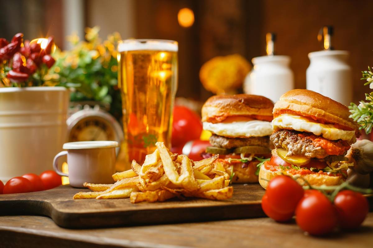 Burgers, fries & beers sit on a wooden table surrounded by tomatoes and some condiments.