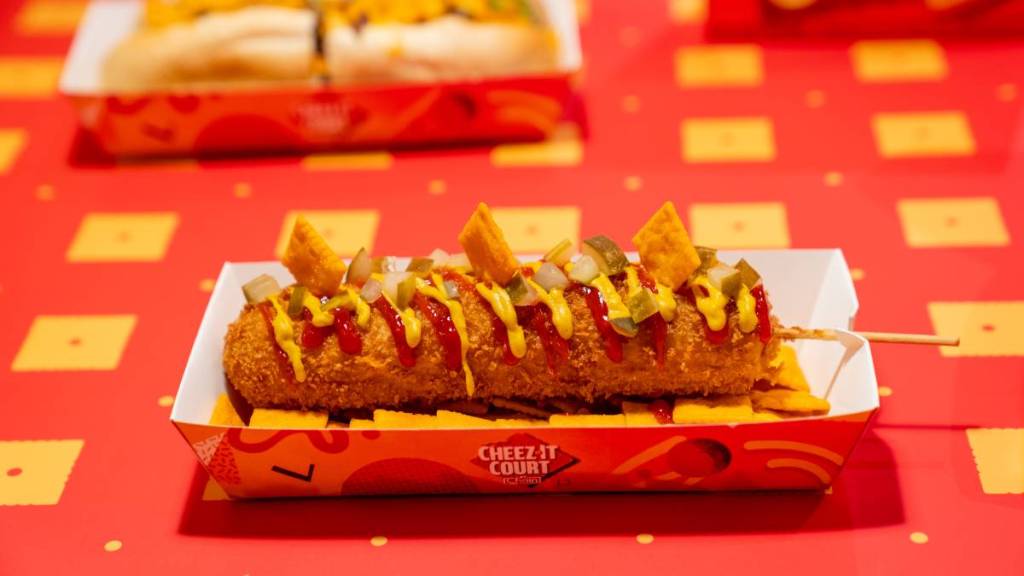 a cheez-it crunchy cheez-dog in a takeout box on a red and yellow tablecloth