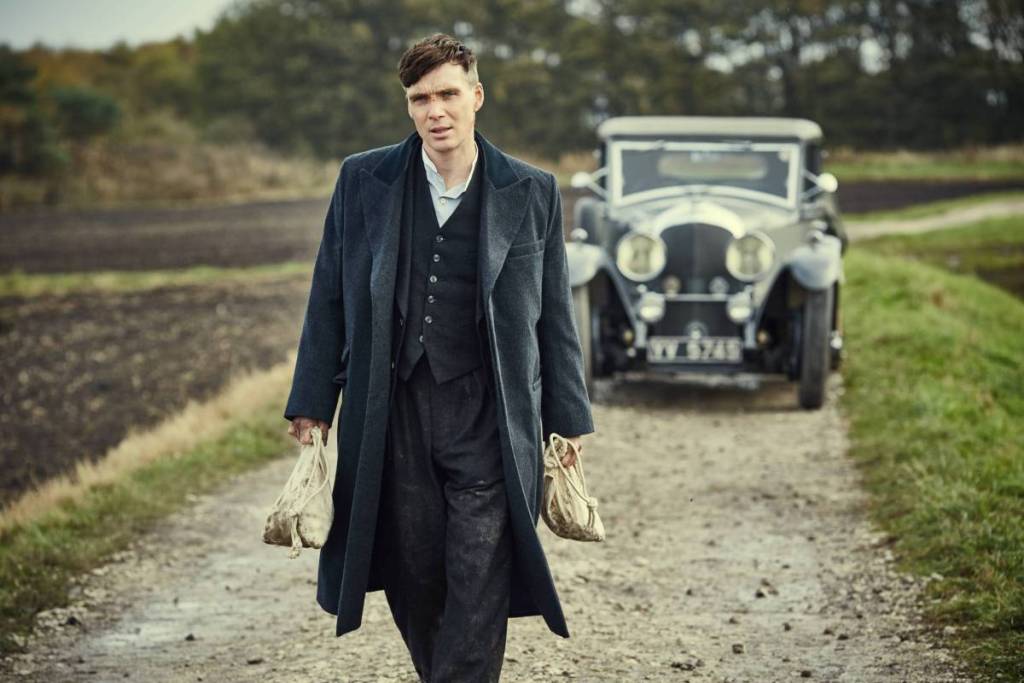 Tommy Shelby spaziert durch die Landschaft und hält zwei Geldsäcke in den Händen