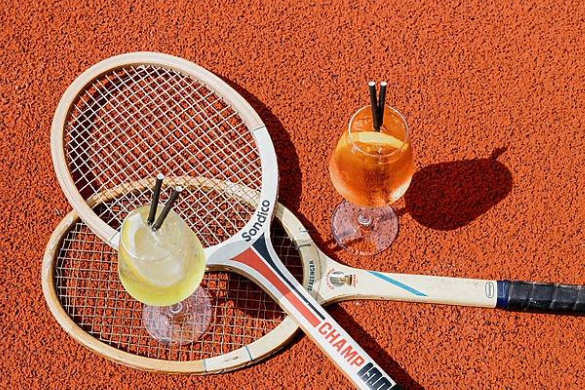 two tennis rackets on courts club manchester top of each other lying on terracotta-coloured court with two cocktails places either side of rackets, one limoncello spritz, the other aperol spritz