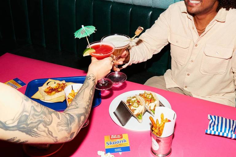 two people cheersing with cocktail classes. there are tacos and fries on the table.