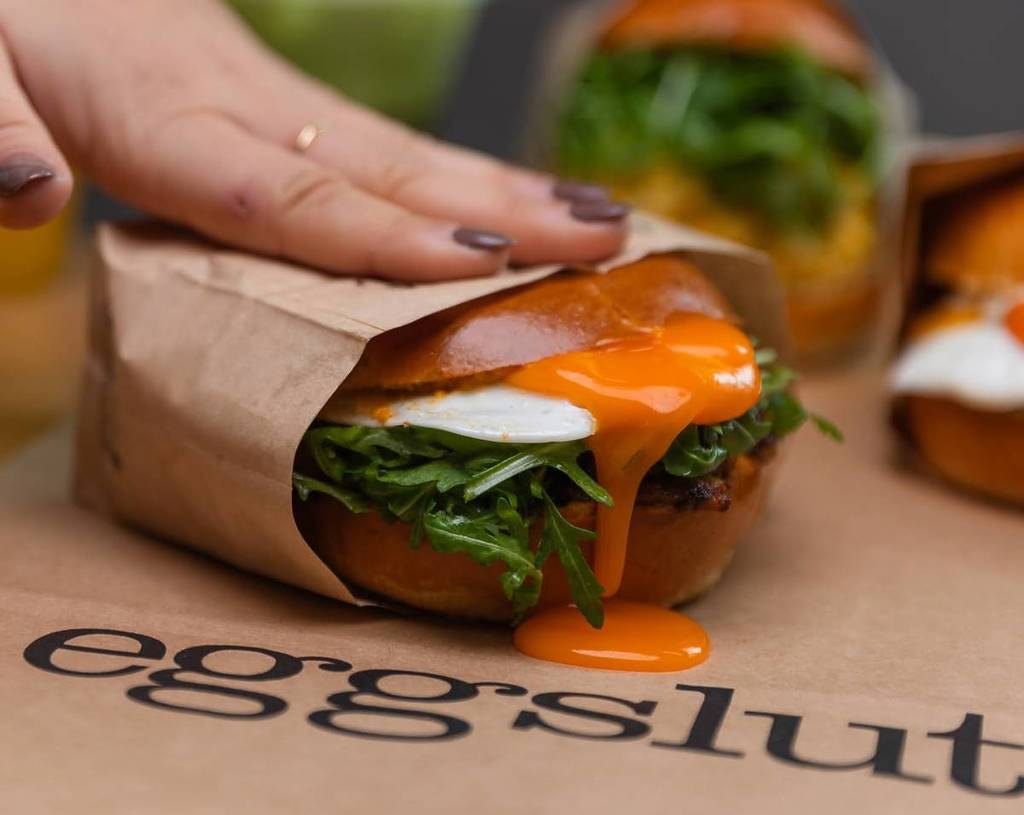 brioche bun containing rocket and fried egg being squished down orange so yolk runs out onto brown paper saying eggslut