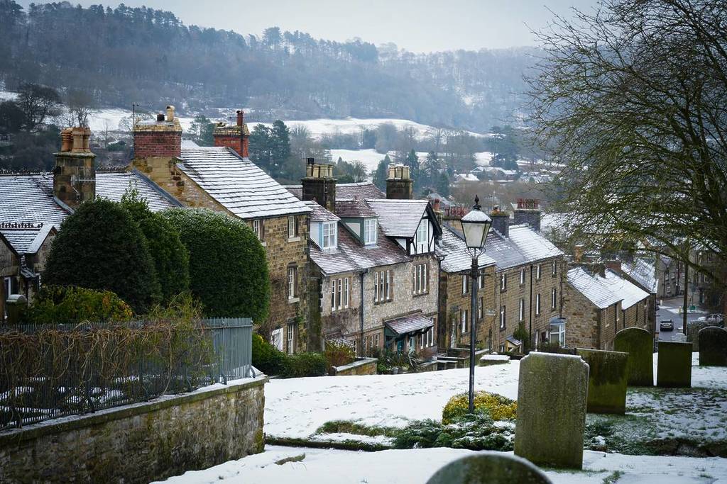 Bakewell, Peak District, UK - January 9, 2025: Snow-covered rooftops of traditional stone cottages in Bakewell, surrounded by bare winter trees and a scenic hillside in the background.