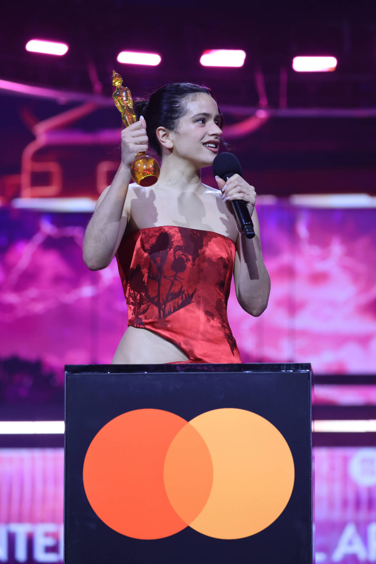 Rosalia in red dress at brit awards in manchester holding microphone and award on podium