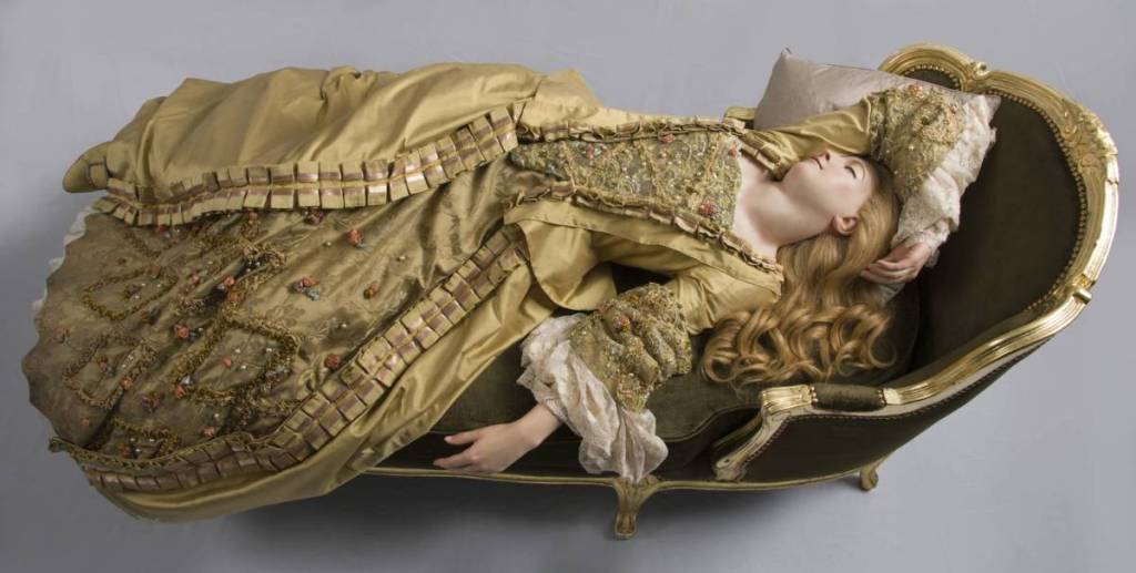 a waxwork figure of a woman sleeping on an ottoman, wearing a golden period-style dress