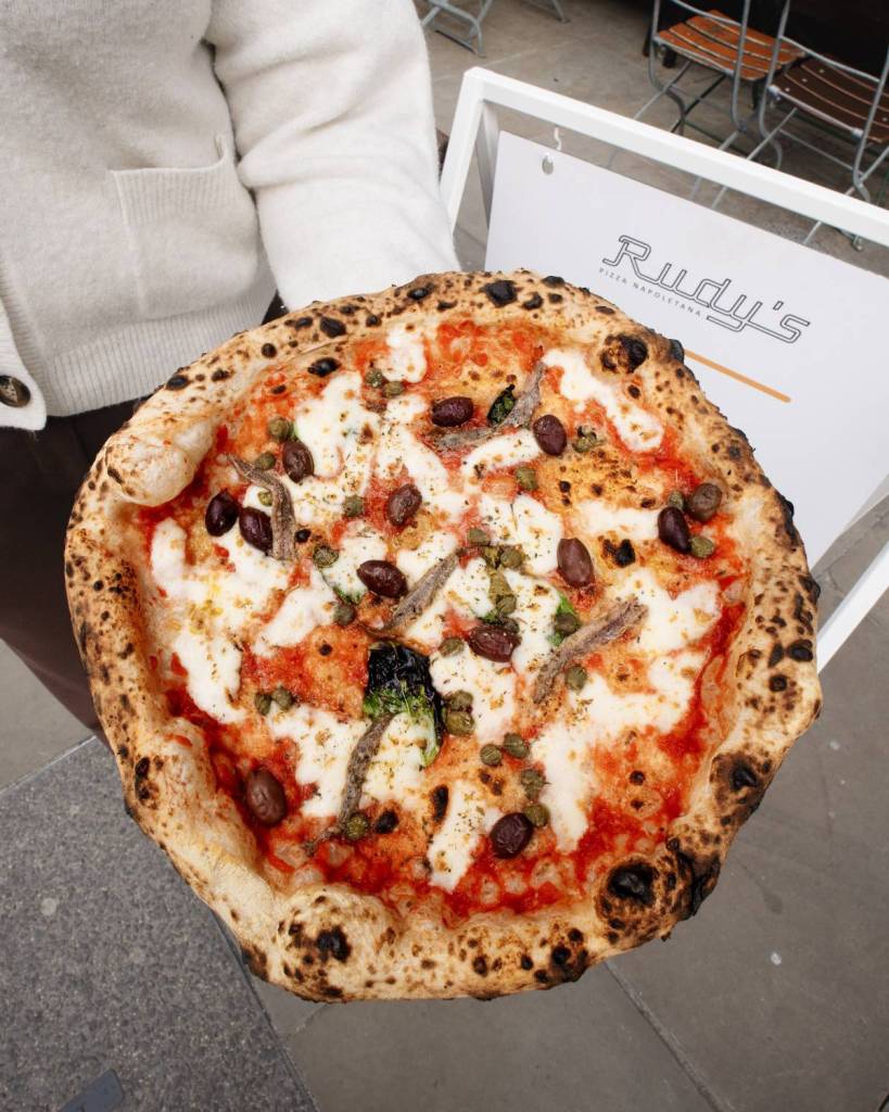 pizza with olives and anchovies on being held outside rudy's sign
