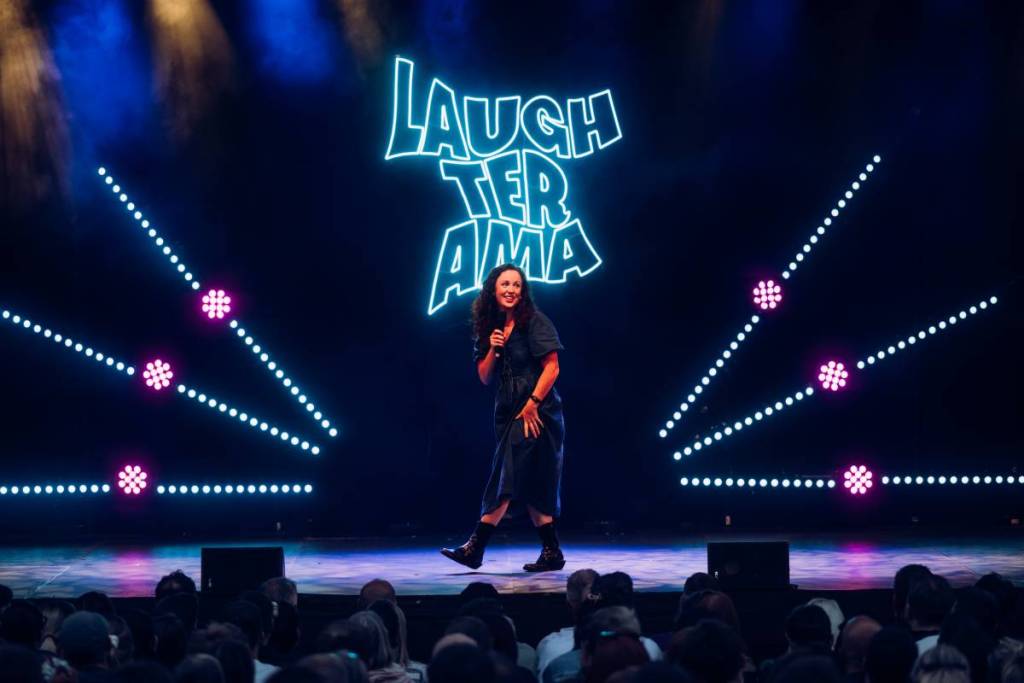 comedian on stage at laughterama 2025 at castlefield bowl, manchester with neon blue laughterama sign behind her