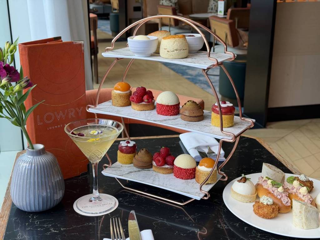 the lowry afternoon tea on circular tier with orange menu and cocktail on left of table and plate of sweets to right of tier