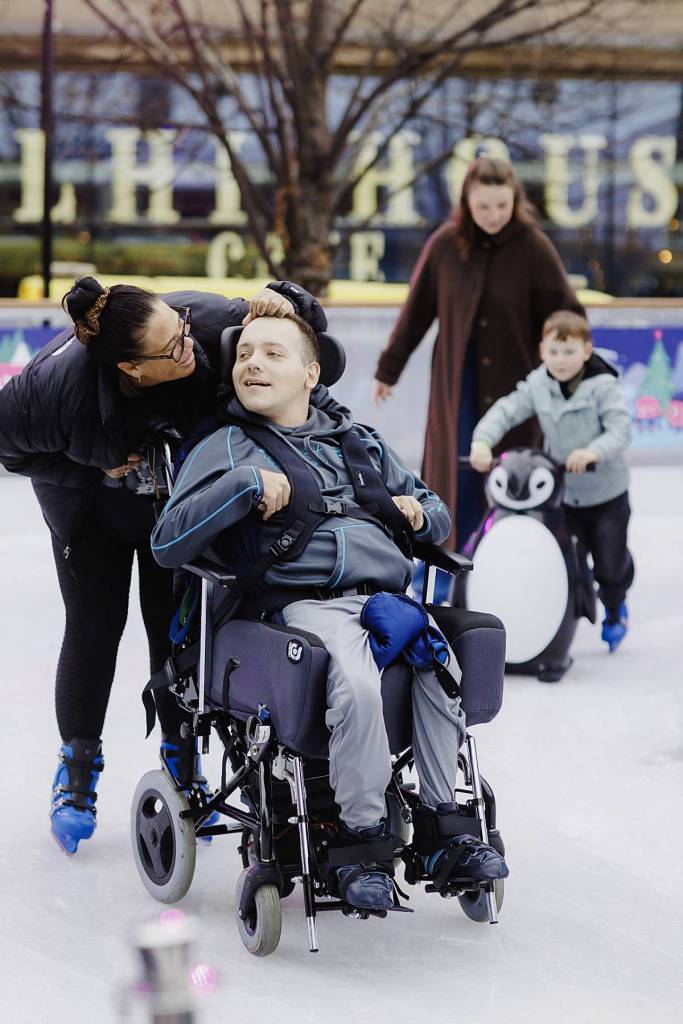 carer pushing young person in wheelchair on skate manchester ice rink and mother and son in background skating with boy pushing penguin skate aid