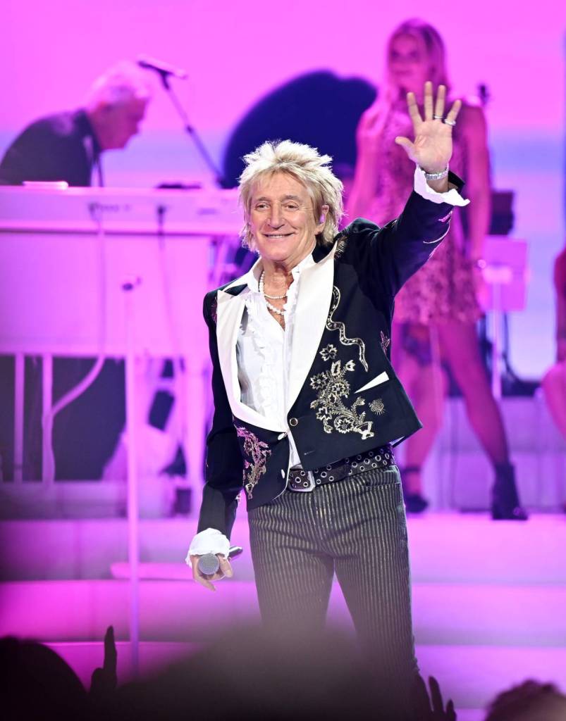 rod stewart wearing black embroidered jacket on stage and waving at crowd with pink lights in background