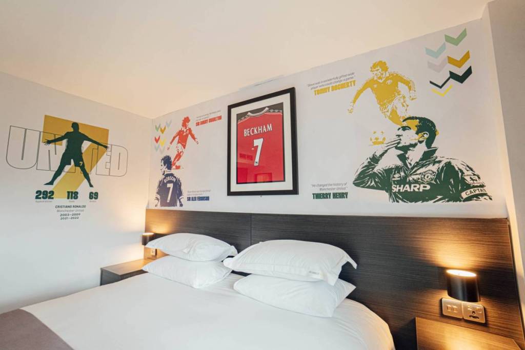 shot of double bed with lights on either side and illustrations of no7 players on walls and david beckham united shirt in frame above bed