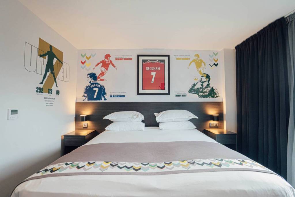shot of double bed in hotel football room with framed david beckham manchester united shirt on wall above bed and illustrations of number seven players on walls