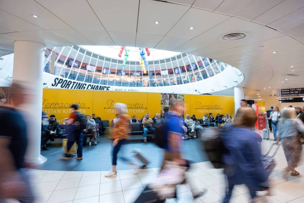 people walking through manchester airport terminal 3 departure lounge with sports abr above