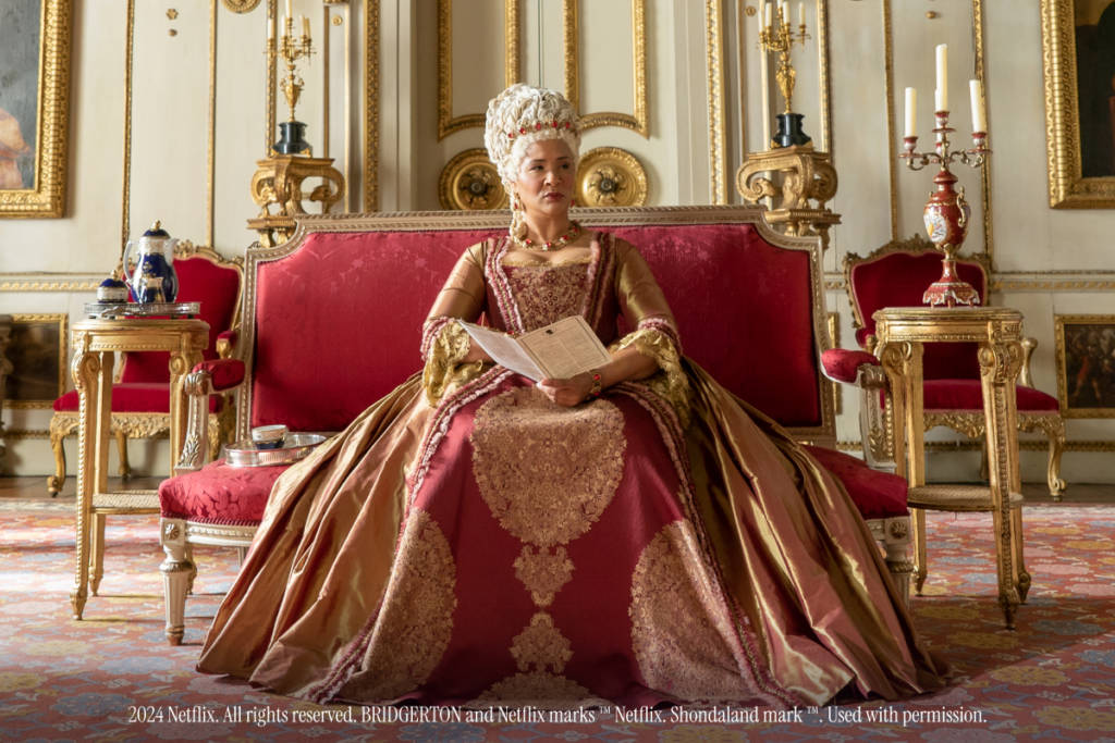 La Reina Carlota, personaje de Los Bridgerton, sentada sobre un sofá rojo de época.