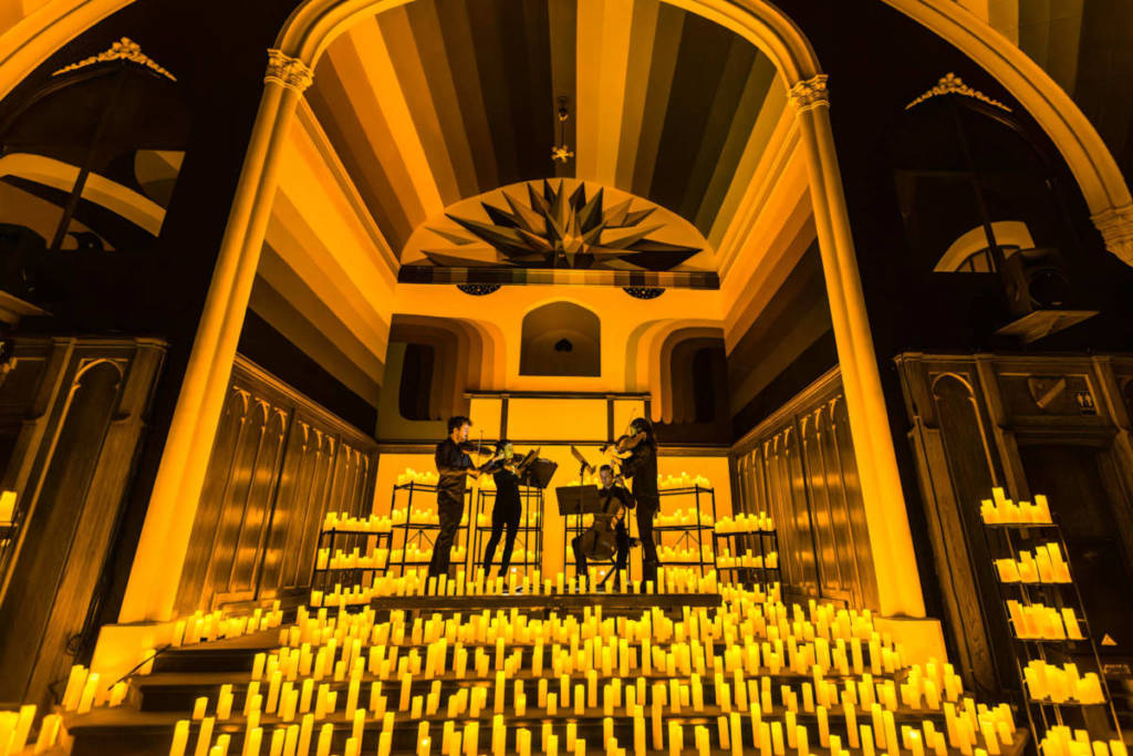 Un cuarteto de cuerda toca durante un concierto Candlelight sobre un escenario cubierto de cálidas velas.