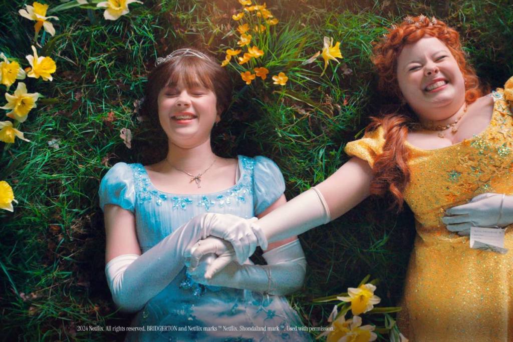 Two characters from the Netflix series Bridgerton laughing and holding hands as they lay on a meadow