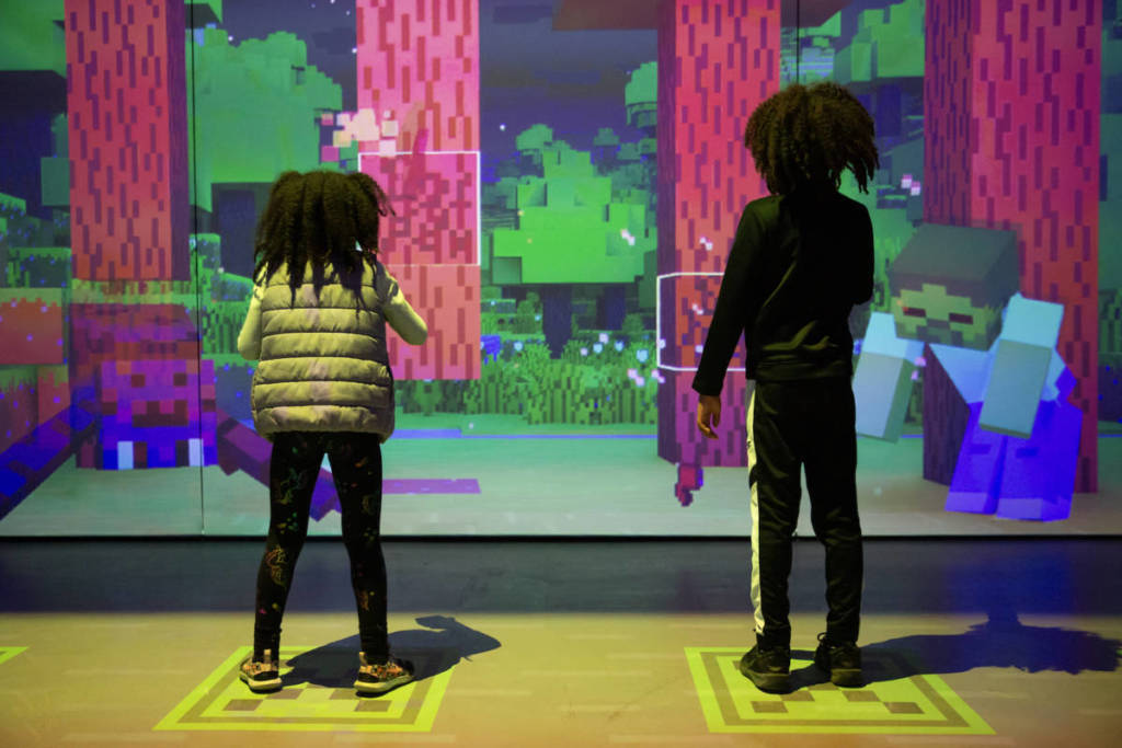 two kids interact with a screen depicting a forest biome from Minecraft