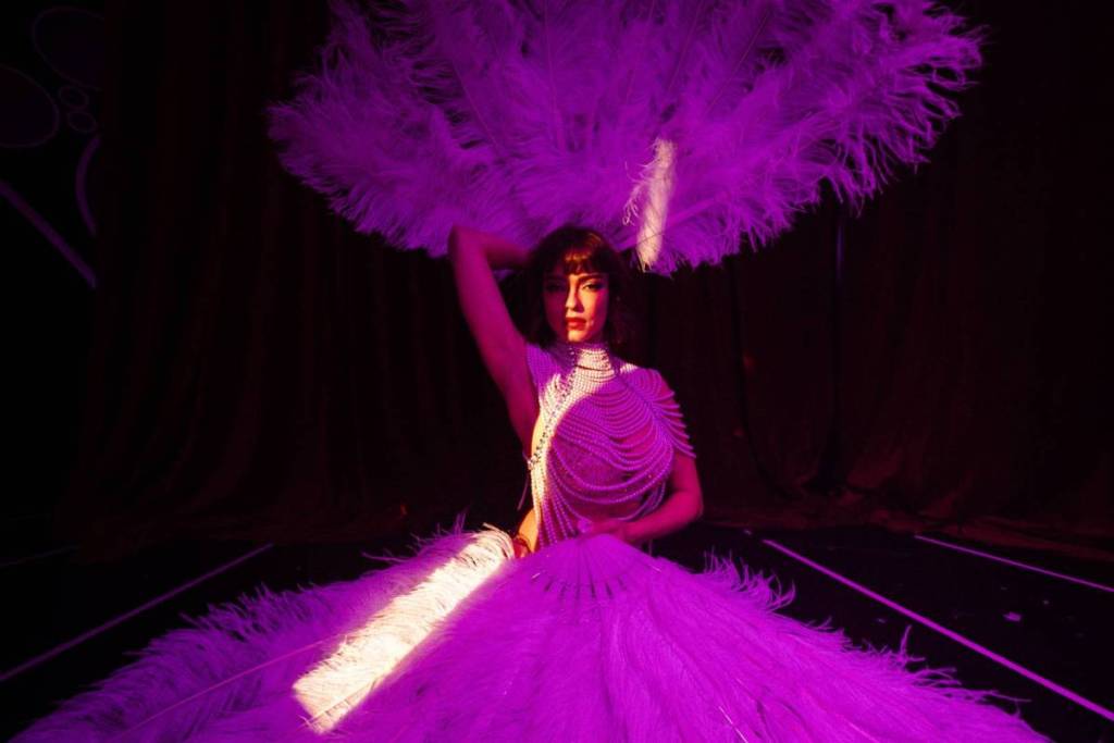 A performer at Loca Vida holds a giant purple feathered wing above and below her.