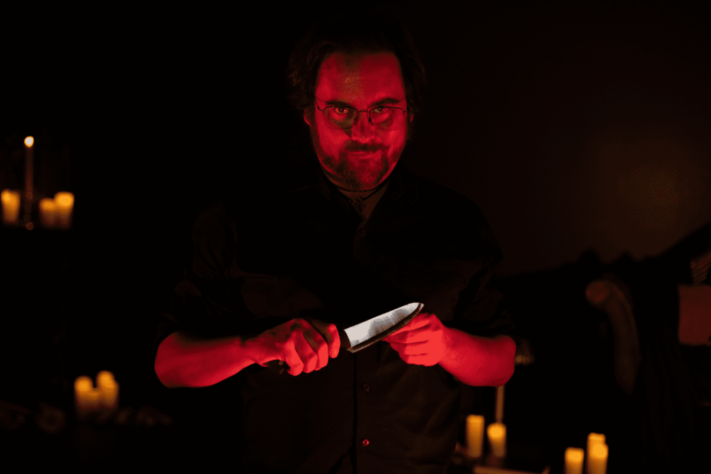 Hombre con gafas sosteniendo un cuchillo en un ambiente oscuro rodeado de velas.